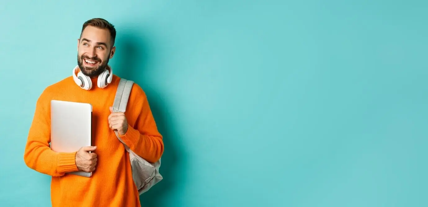 Fröhlicher Mann mit Rucksack und Kopfhörern, der einen Laptop hält, lächelnd und nachdenklich nach links blickend, stehend.